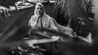 Siyah beyaz klasik fotoğraf, genç sarışın, pencerenin yanındaki sandalyede oturan güzel kız tropik çiçekli beyaz gömlek, organik cilt bakımı, lüks spa oteli, yaşam tarzı fotoğrafı. Seçici odak