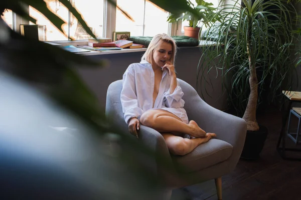 Genç sarışın, pencerenin yanındaki sandalyede oturan güzel bir kız tropik çiçekli beyaz bir gömlek, organik cilt bakımı, lüks bir spa oteli, yaşam tarzı fotoğrafı. Seçici odak