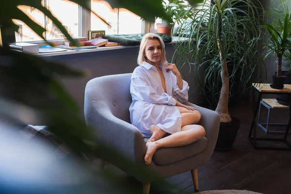 Genç sarışın, pencerenin yanındaki sandalyede oturan güzel bir kız tropik çiçekli beyaz bir gömlek, organik cilt bakımı, lüks bir spa oteli, yaşam tarzı fotoğrafı. Seçici odak