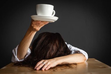 Woman with arms outstretched, head on table, holding cup of coffee or tea, sitting, working at desk, isolated on gray background, tiredness, morning. Business career achievement concept. Copy