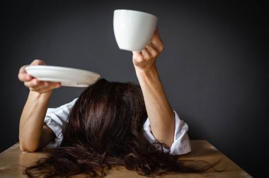 Woman with arms outstretched, head on table, holding cup of coffee or tea, sitting, working at desk, isolated on gray background, tiredness, morning. Business career achievement concept. Copy
