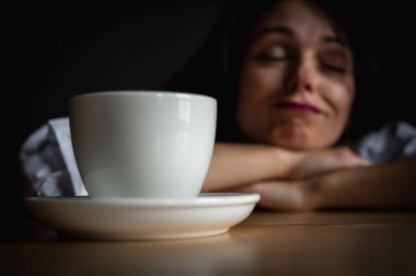 Yakın plan portre. Elleriyle masaya yaslanan genç ve çekici bir kız yorgun, bitkin bir şekilde sabah kahvesinin yanında yüzünü gösteriyor. Zor bir gün, karanlık bir geçmiş. seçici odak