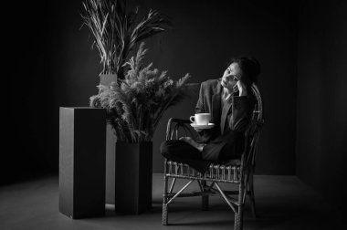 Black and white classic photo. Morning rest. Young beautiful girl sits in a wooden wicker chair and enjoys coffee near the window, free space. Selective focus, grain