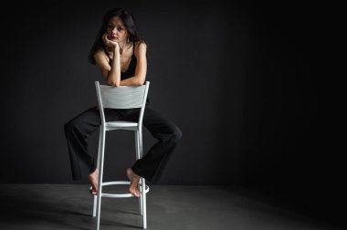 Güzel ve baştan çıkarıcı esmer bir kadının tam vücut fotoğrafı. Koyu arka planda izole edilmiş bir sandalyede pantolonlu oturuyor. 