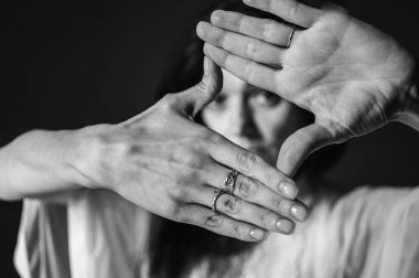 Endişe, tehlike, koruma. Bir kadının yüzünü elleriyle örttüğü kavramsal görüntü. Gözleriyle karanlık bir stüdyo arka planında parmaklarının arasından bakıyor.