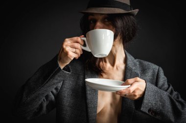 Şapkalı ve gri ceketli bir kız elinde sabah kahvesi ya da gri arka planda çay olan beyaz bir bardak tutuyor. Seçici odak, arkaplanda izole