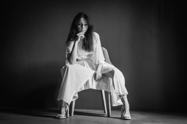 Siyah beyaz klasik fotoğraf, yan odak. Genç, çekici, düşünceli, pozitif, güzel bir kızın fotoğrafı, eller, yanaklar, sandalyede oturuyor, açık renk elbise giyiyor, gri arka planda izole edilmiş.