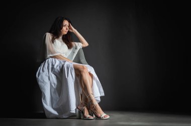 Genç, çekici, düşünceli, pozitif, güzel bir kızın fotoğrafı, eller, yanaklar, sandalyede oturuyor, açık renk elbise giyiyor, gri arka planda izole edilmiş.