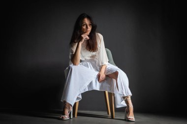 Genç, çekici, düşünceli, pozitif, güzel bir kızın fotoğrafı, eller, yanaklar, sandalyede oturuyor, açık renk elbise giyiyor, gri arka planda izole edilmiş.