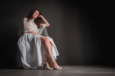 Genç, çekici, düşünceli, pozitif, güzel bir kızın fotoğrafı, eller, yanaklar, sandalyede oturuyor, açık renk elbise giyiyor, gri arka planda izole edilmiş.