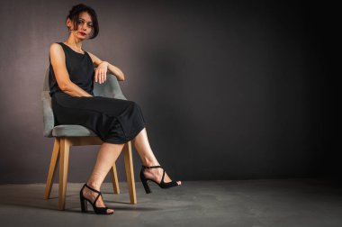 Siyah elbiseli, yorgun, işkence görmüş, genç ve şehvetli bir kadının stüdyo portresi. Tahta bir sandalyeye oturmuş, arkasına yaslanmış, karanlık bir arka planda. Depresyon