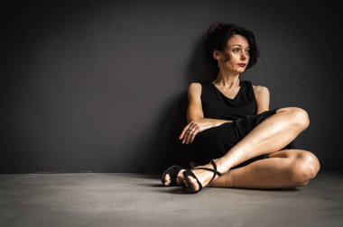 Yorgun, işkence görmüş, siyah elbiseli, gri arka planda yerde oturan genç ve şehvetli bir kadının stüdyo portresi. Depresyon, yorgunluk, yalnızlık, düşüncelilik.