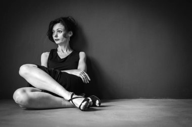 Yorgun, işkence görmüş, siyah elbiseli, gri arka planda yerde oturan genç ve şehvetli bir kadının stüdyo portresi. Depresyon, yorgunluk, yalnızlık, düşüncelilik. Siyah ve beyaz