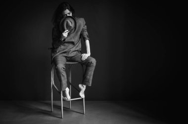 B & W, zarif, güzel, gri takım elbiseli, erkek şapkası takan, sandalyede oturan seksi bir kadının yakın plan portresi. Koyu gri arka planda kırmızı dudaklı ve göz makyajlı güzel bir manken.