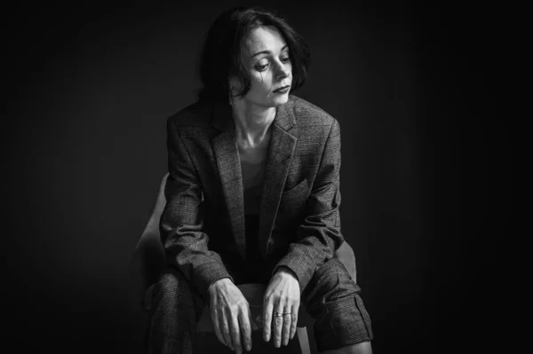 B&W, Close-up portrait of an elegant, pretty, sexy woman in a gray suit, wearing a man's hat, sitting on a chair. Beauty fashion model with red lips and eye makeup on a dark gray background