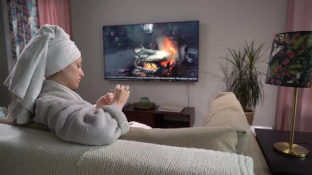 Μια νεαρή γυναίκα κάνει μανικιούρ καθισμένη στον καναπέ στο σαλόνι με τηλεόραση. — Αρχείο Βίντεο