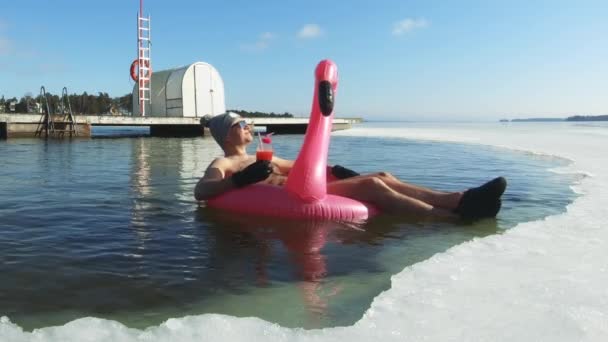 Ein Mann träumt von einem Urlaub, der auf einem aufblasbaren Flamingo in einem Eisloch schwimmt — Stockvideo