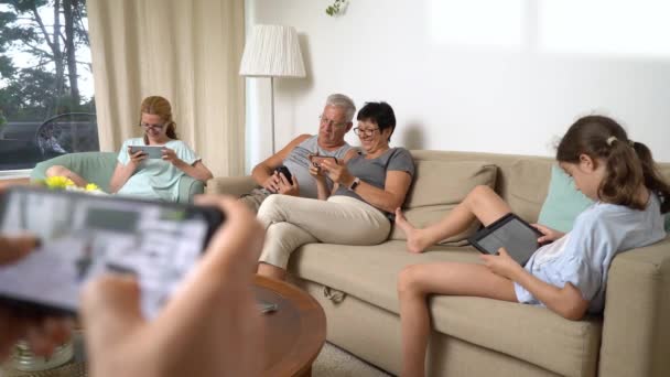 大きな幸せな家族が一緒に自宅でオンラインモバイルゲームをプレイしている 人々はソファの上に座って楽しんでいるさまざまな年齢層 両親祖父母と少女モバイルデバイスでリラックスしたリビングルームで — ストック動画