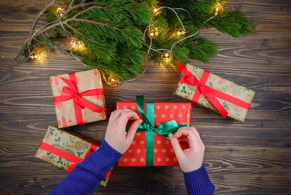 Noel hediyelerini ahşap arka planda Noel ağacı ve yılbaşı ışıkları dallarıyla paketliyor.