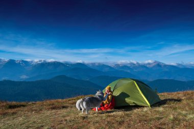 Sibirya köpeğiyle birlikte kamp çadırının yanında oturan genç bir kız. Arka planda dağların zirveleri ve mavi gökyüzü var. İnsan ve hayvan arkadaşlığı kavramı