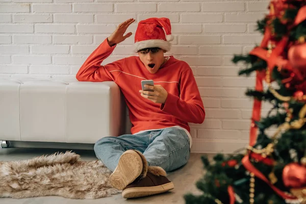 クリスマスに家にいる若い男が携帯電話を見て驚いて — ストック写真