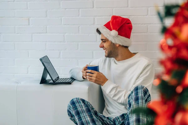 サンタ クラスの帽子を被った男がクリスマスにラップトップを持って家に — ストック写真