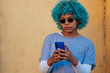 Cep telefonu ve mavi saçlı güneş gözlüklü Afro-Amerikan kız portresi.