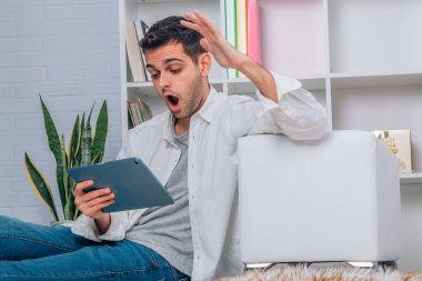 Evde tablet veya dizüstü bilgisayarı olan ve şaşırmış ifadesi olan bir adam.