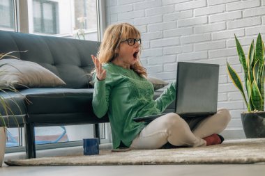 Evde bilgisayar ya da dizüstü bilgisayara bakan şaşkın ve şok olmuş bir kadın.