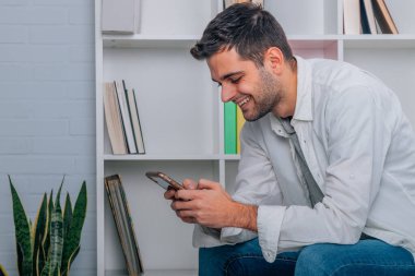 Evdeki genç adam koltukta oturuyor ve cep telefonuna bakıyor.