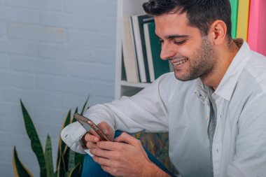 Evdeki genç adam koltukta oturuyor ve cep telefonuna bakıyor.