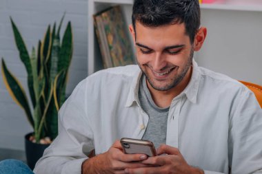 Evdeki genç adam koltukta oturuyor ve cep telefonuna bakıyor.