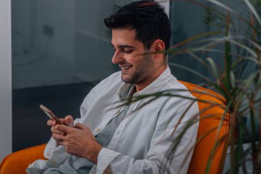 Evdeki genç adam koltukta oturuyor ve cep telefonuna bakıyor.