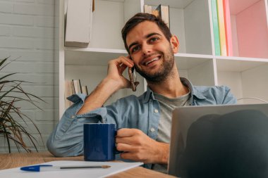 Adam evde bilgisayarla çalışıyor ve cep telefonu kullanıyor.