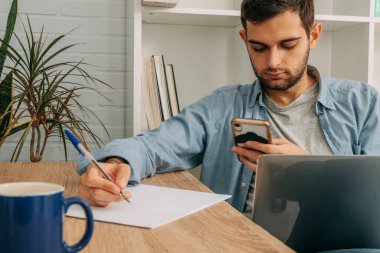 Bilgisayarlı, cep telefonlu ya da akıllı telefonlu genç adam not alıyor