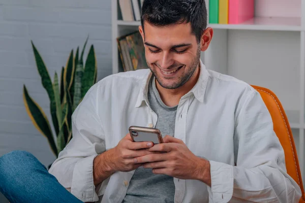Evdeki genç adam koltukta oturuyor ve cep telefonuna bakıyor.