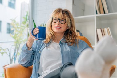 Orta yaşlı bir kadın evde rahatça kahve içiyor.