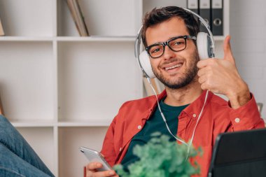 Evde cep telefonu ve kulaklıkla müzik dinleyen genç bir adam var.