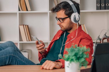 Evde cep telefonu ve kulaklıkla müzik dinleyen genç bir adam var.