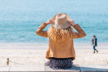 Kumsalda kumsalda oturan şapkalı kız.