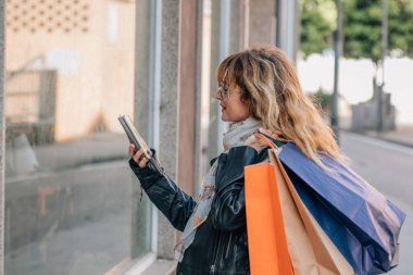 Alışveriş torbaları ve cep telefonuyla vitrinlere bakan orta yaşlı bir kadın.