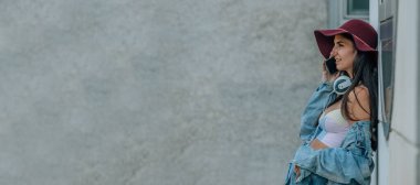 Şapkalı kız sokakta duvarda cep telefonuyla konuşuyor.