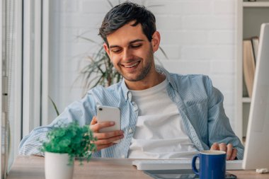 Evdeki genç adam cep telefonu ve bilgisayarla çalışıyor.