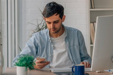 Evdeki genç adam cep telefonu ve bilgisayarla çalışıyor.