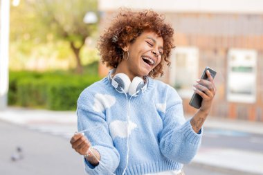 Gülümseyen kız cep telefonuna bakıyor ve kulaklık kablosuyla oynuyor.