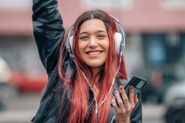 Kızıl saçlı kız mutlu gülümsüyor, cep telefonu ve kulaklıkla sokakta.