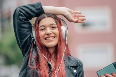 Kızıl saçlı kız mutlu gülümsüyor, cep telefonu ve kulaklıkla sokakta.