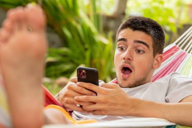 Yazın hamakta cep telefonu ve sürpriz ifadesiyle genç bir adam.