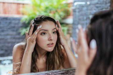 Banyoda aynanın karşısında kız parmaklarıyla yüzüne dokunur ve cildine bakar. Sivilce ya da kırışıklık arar.