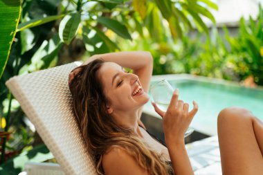 Havuzun arka planında ve yeşilliklerin üzerinde elinde bardakla gülümseyen geniş bir kız oturuyor.